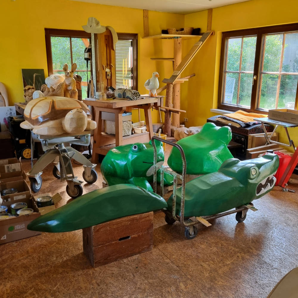 Bemalte Krokodilskulptur aus Holz in der Werkstatt mit weiteren Tierskulpturen im Hintergrund.