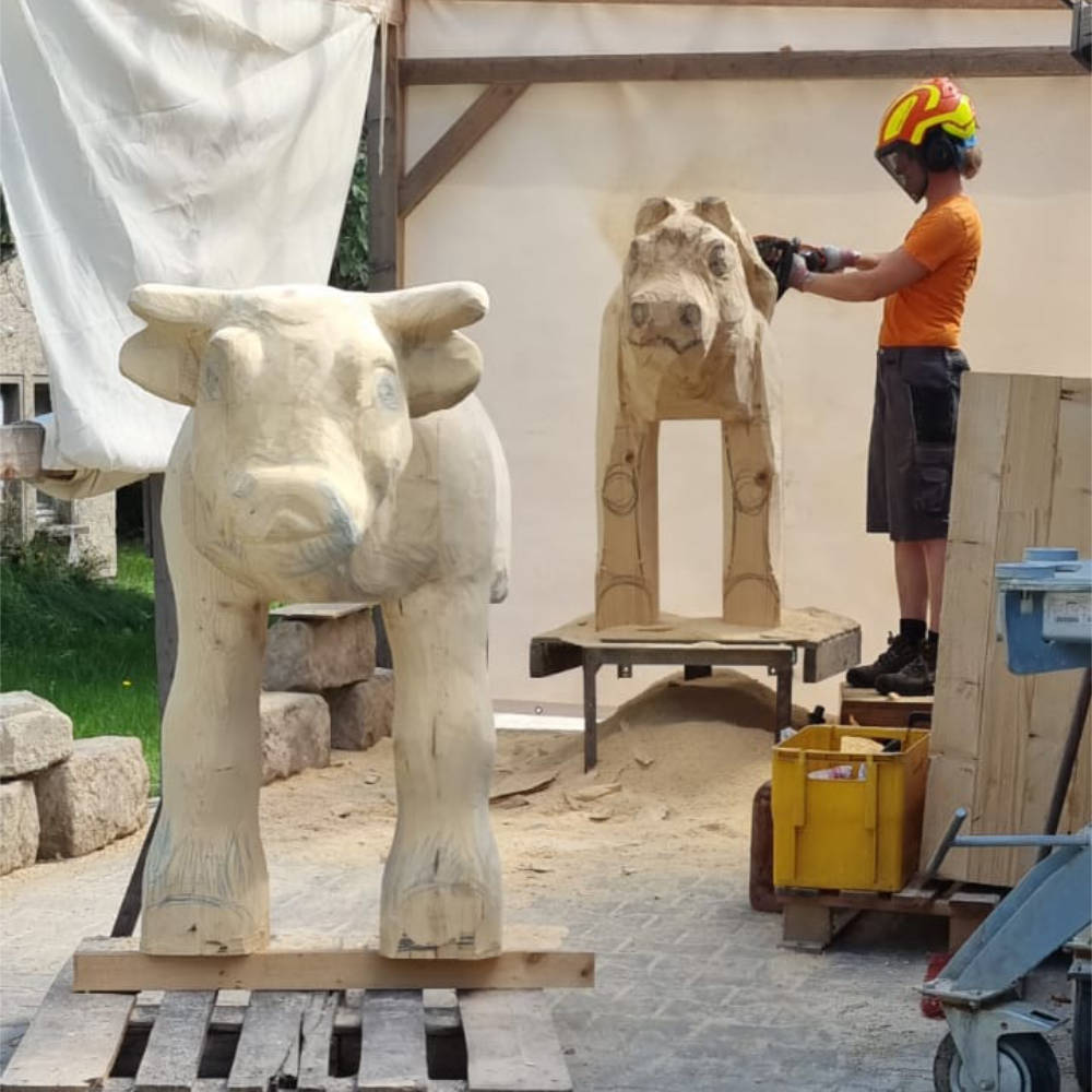 Fein ausgearbeitete Kuhskulptur aus Holz mit Juliane Uebe im Hintergrund bei der Bearbeitung einer weiteren Skulptur. 