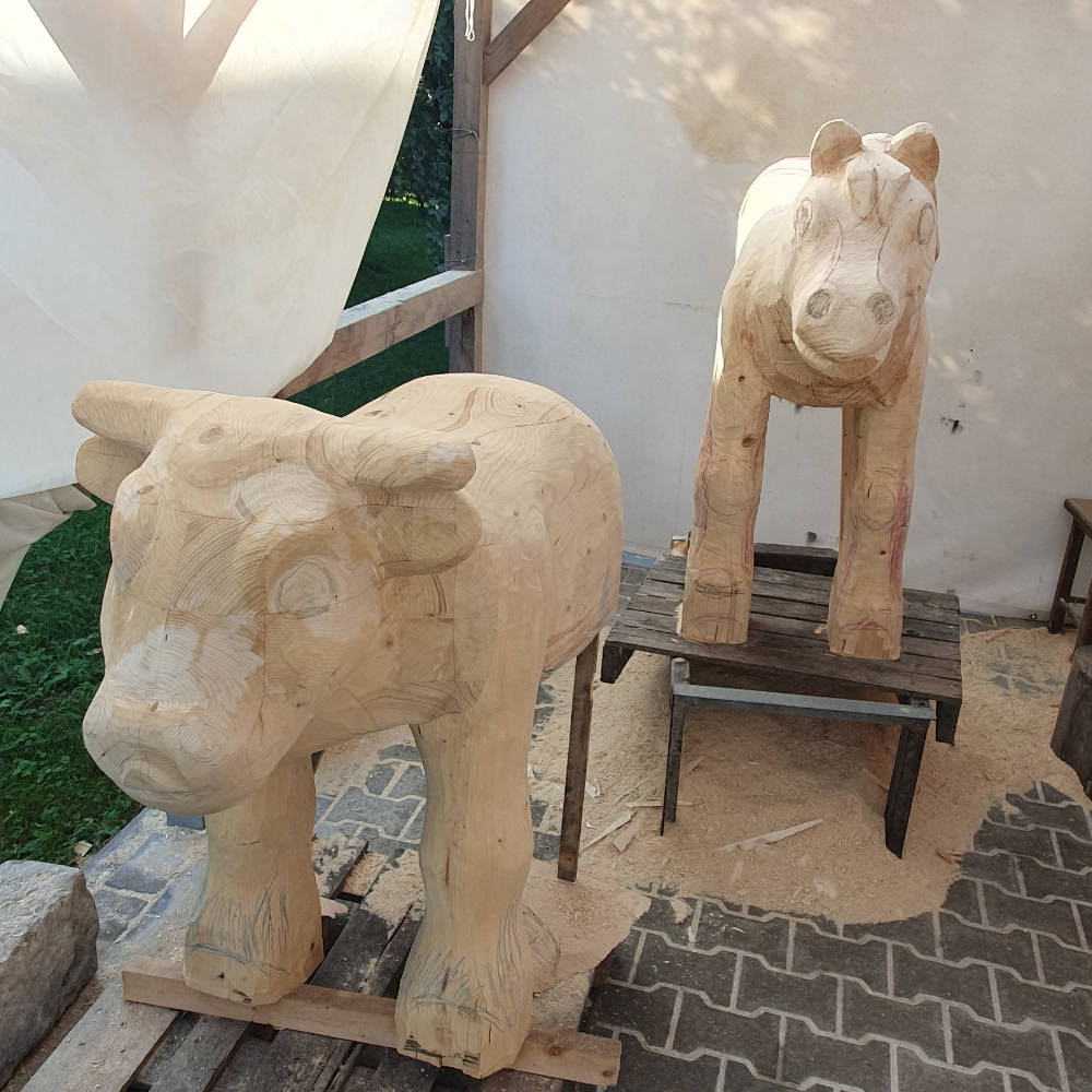 Fertige, unbemalte Kuhskulptur aus Holz mit einer Pferdeskulptur im Hintergrund.