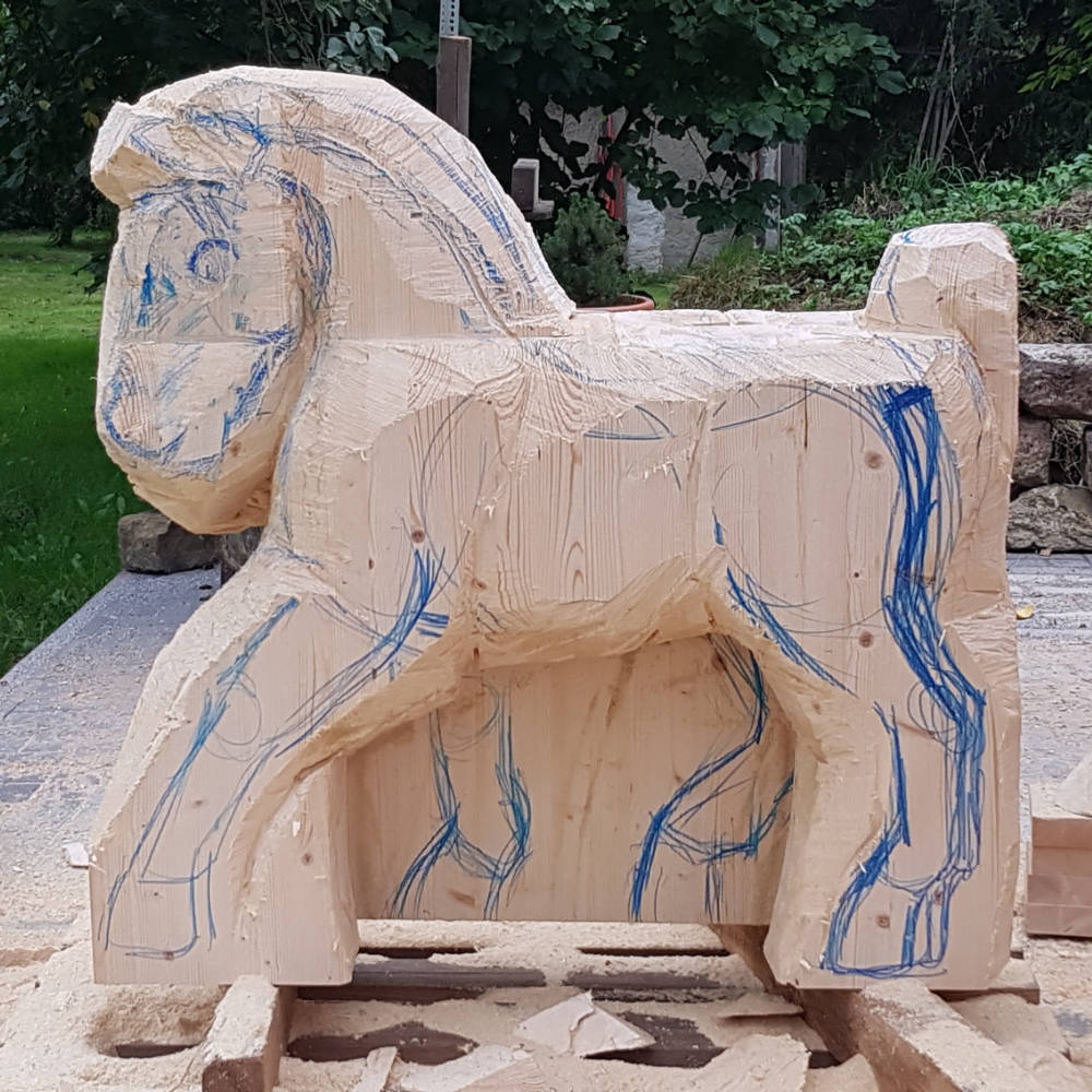 Grob zugesägter Holzblock in Pferdeform mit angezeichneten Konturen für Beine und Kopfbereich.