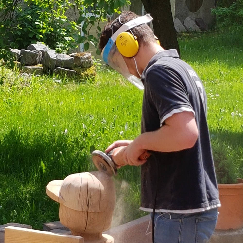 Mann bearbeitet Pferdeskulptur aus Holz mit einer Bohrmaschine, während er auf der Figur sitzt.